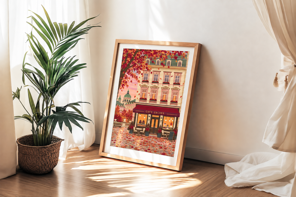 Framed cross stitch featuring an Austrian cafe storefront illustration with autumn leaves, a beige building facade, and a warm pink sky, shown in a light wood frame against a white wall.