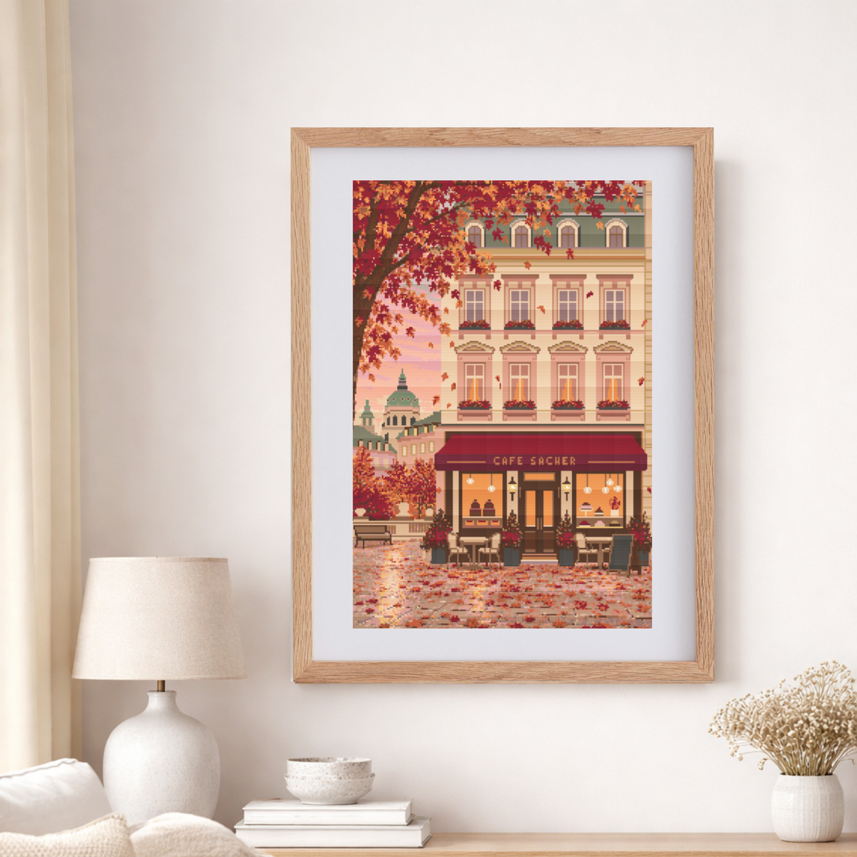Framed cross stitch featuring an Austrian cafe storefront illustration with autumn leaves, a beige building facade, and a warm pink sky, shown in a light wood frame against a white wall.