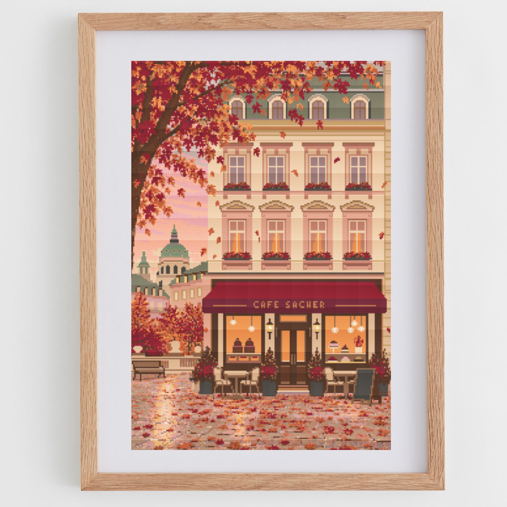 Framed cross stitch featuring an Austrian cafe storefront illustration with autumn leaves, a beige building facade, and a warm pink sky, shown in a light wood frame against a white wall.