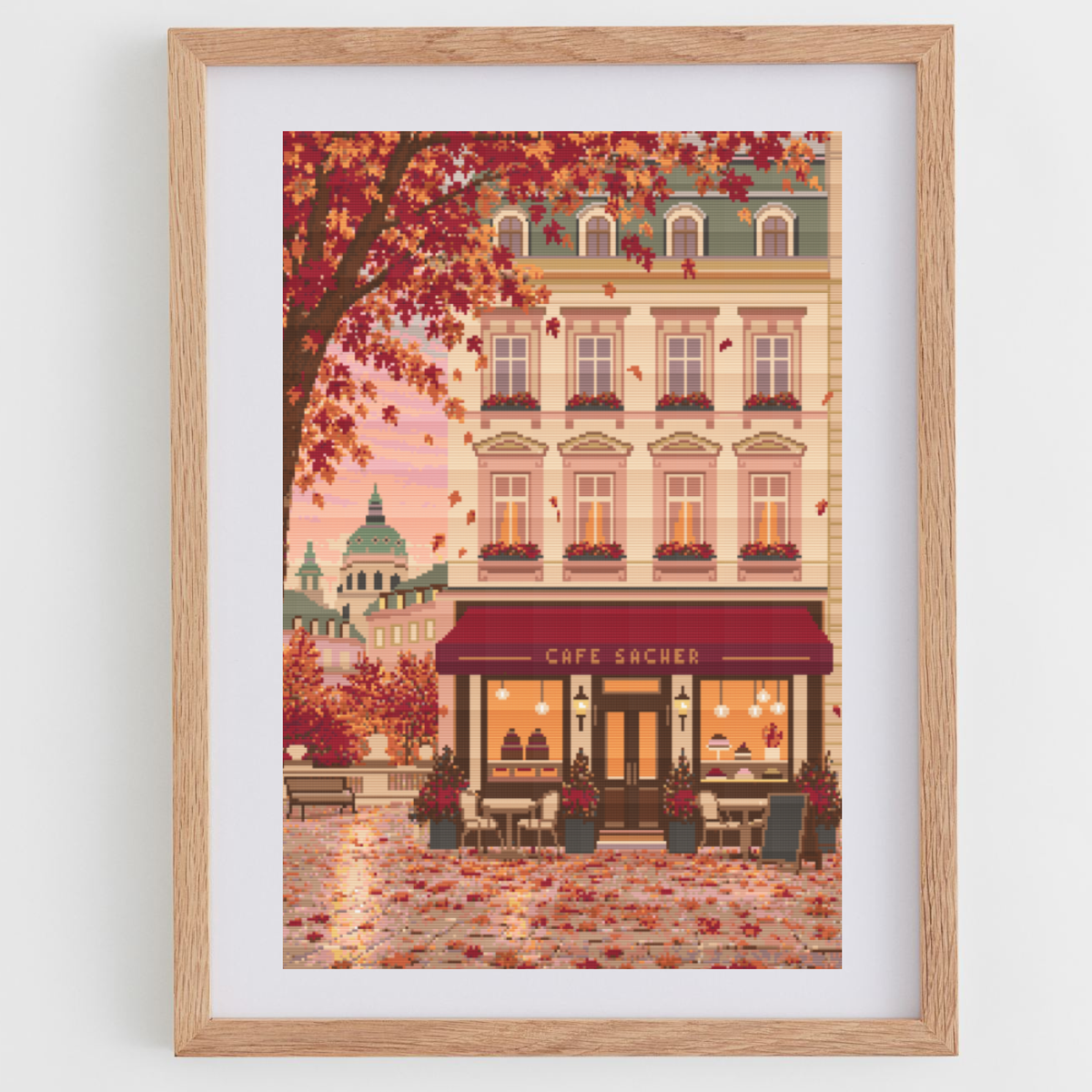Framed cross stitch featuring an Austrian cafe storefront illustration with autumn leaves, a beige building facade, and a warm pink sky, shown in a light wood frame against a white wall.