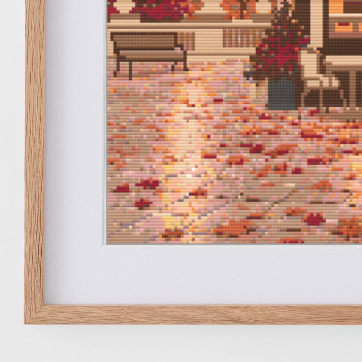 Framed cross stitch featuring an Austrian cafe storefront illustration with autumn leaves, a beige building facade, and a warm pink sky, shown in a light wood frame against a white wall.