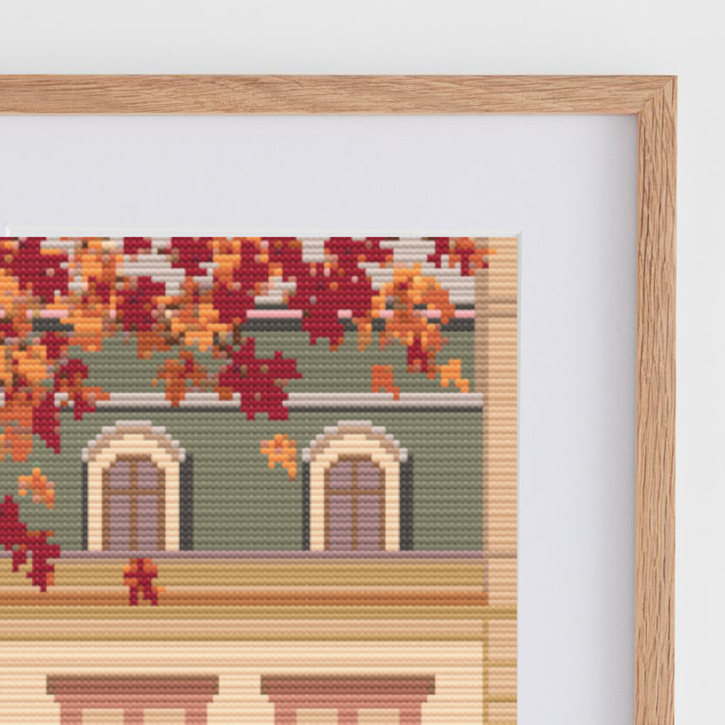 Framed cross stitch featuring an Austrian cafe storefront illustration with autumn leaves, a beige building facade, and a warm pink sky, shown in a light wood frame against a white wall.