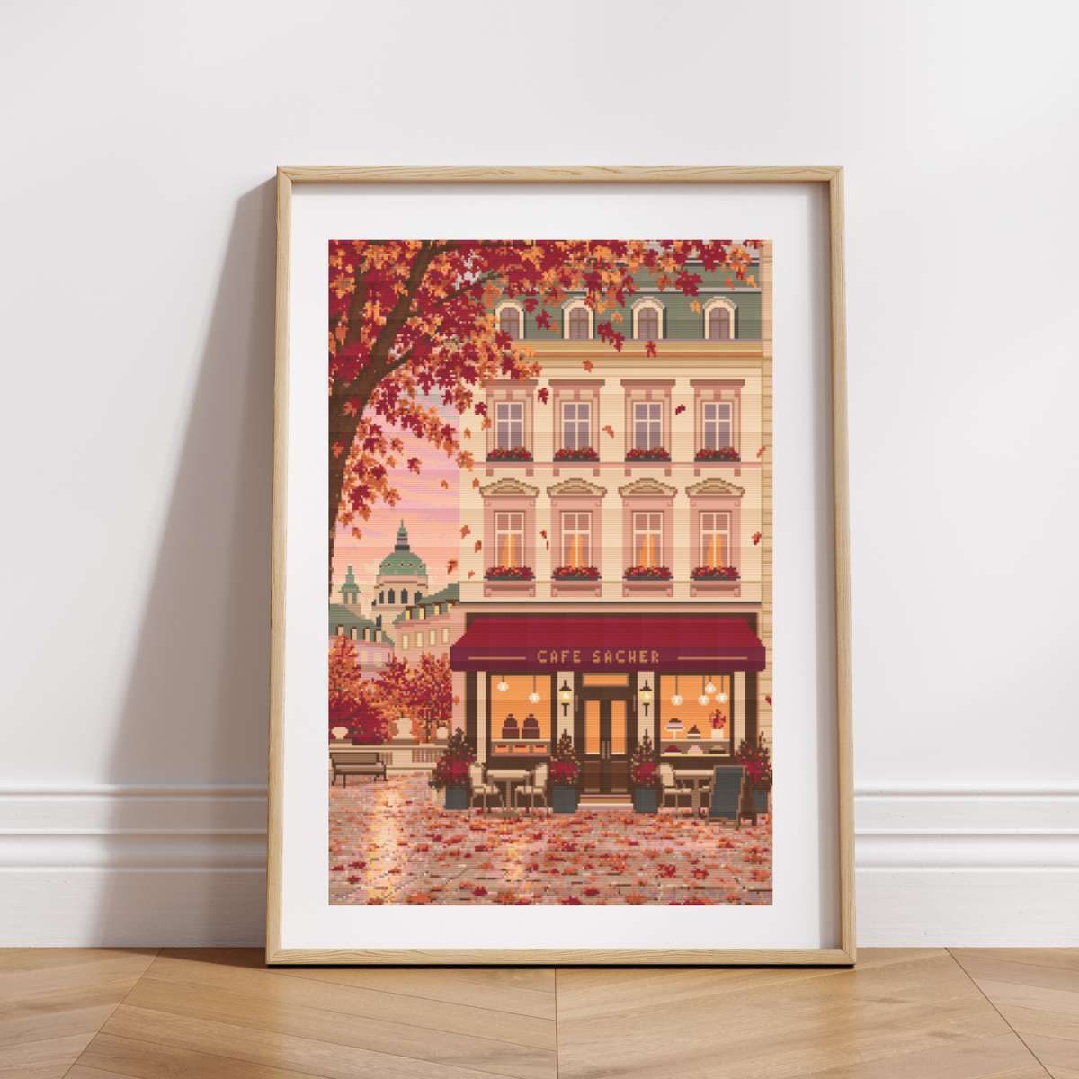 Framed cross stitch featuring an Austrian cafe storefront illustration with autumn leaves, a beige building facade, and a warm pink sky, shown in a light wood frame against a white wall.