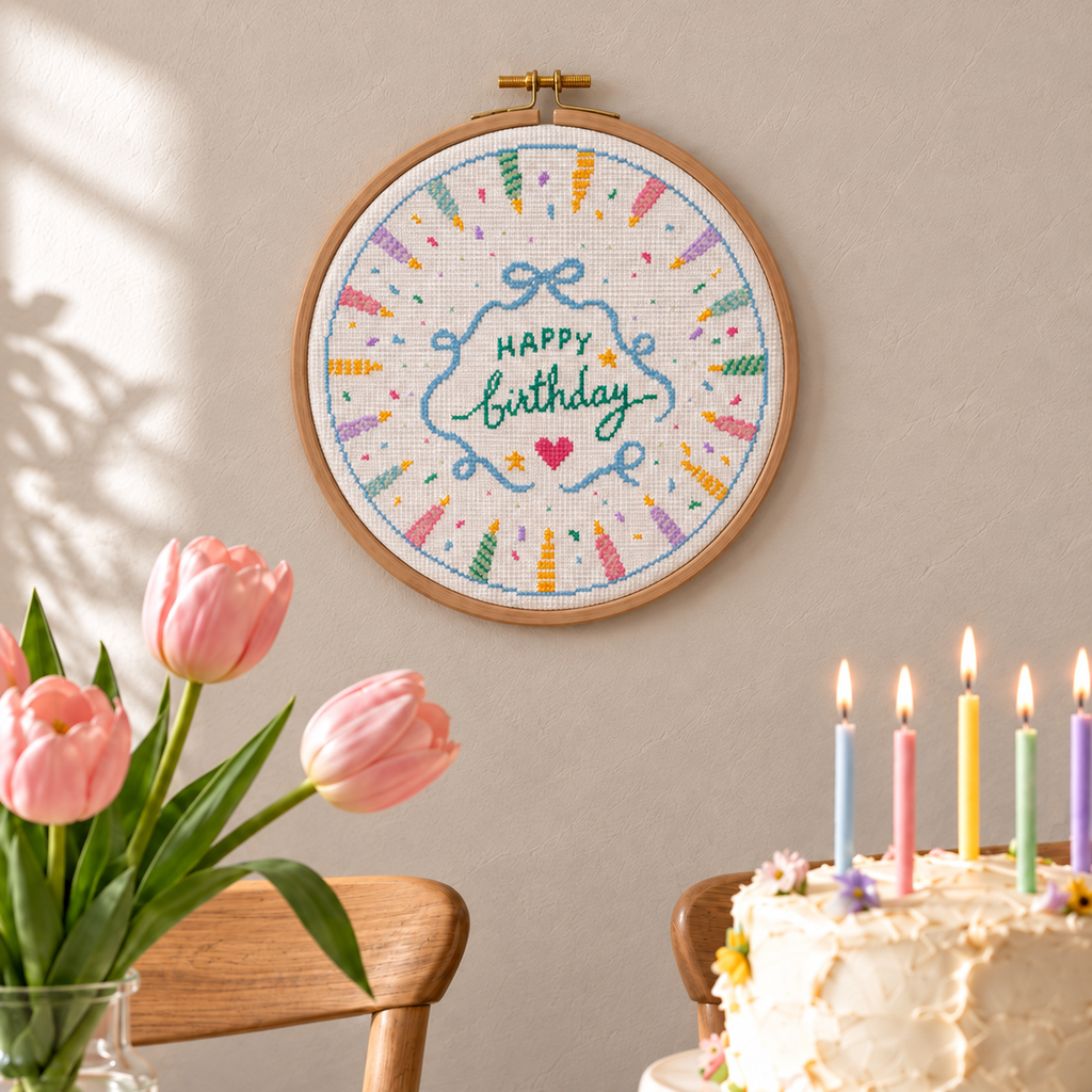 A round wooden embroidery hoop hanging on a beige wall, featuring a colorful cross-stitch design that says 'Happy birthday' with confetti and candles. In the foreground, pink tulips in a vase and part of a birthday cake with lit candles are visible.