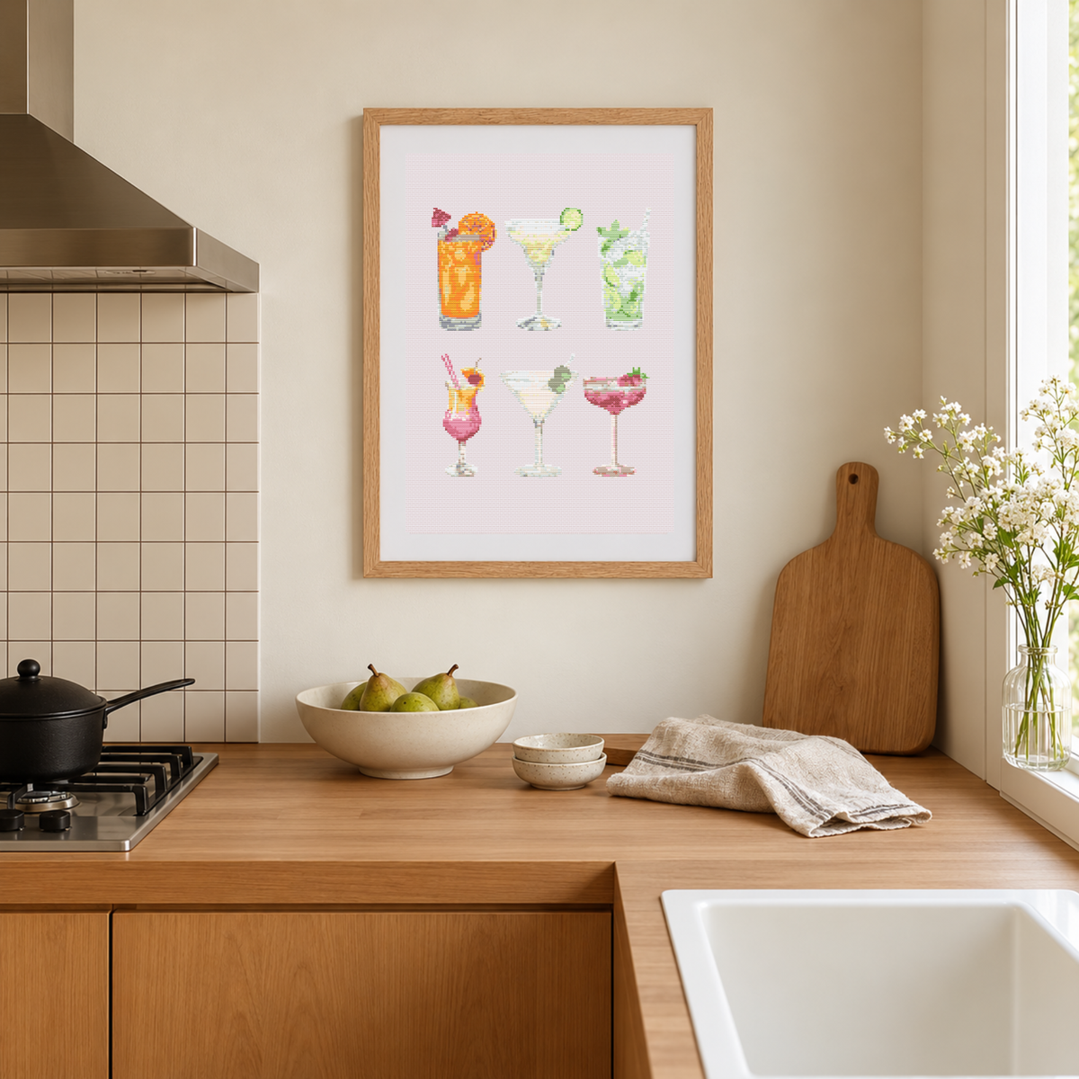 A framed cross-stitch pattern featuring six colorful watercolor cocktails on a pale pink background, hanging on a beige wall in a modern kitchen setting with wooden countertops, a sink, and a window.
