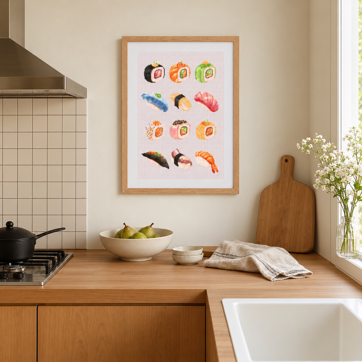 A framed cross-stitch pattern featuring colorful watercolor illustrations of various sushi types, hanging on a beige wall in a modern kitchen setting with wooden countertops, a bowl of pears, and a vase of flowers.