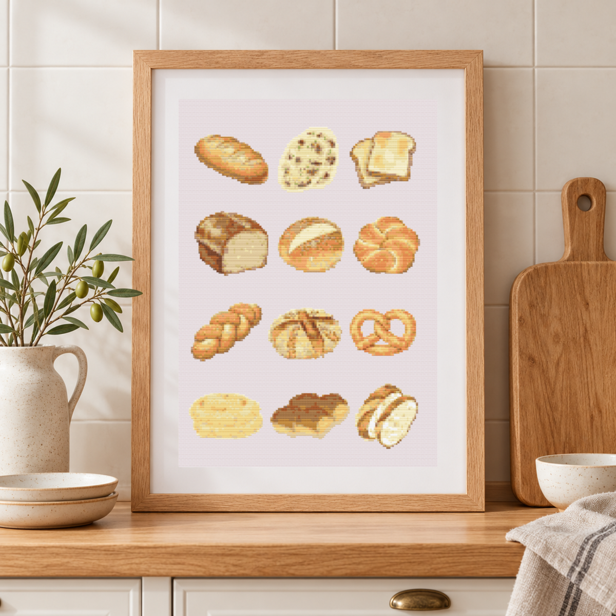 A framed cross-stitch pattern featuring  various breads and pastries, displayed on a beige wall above a kitchen counter.