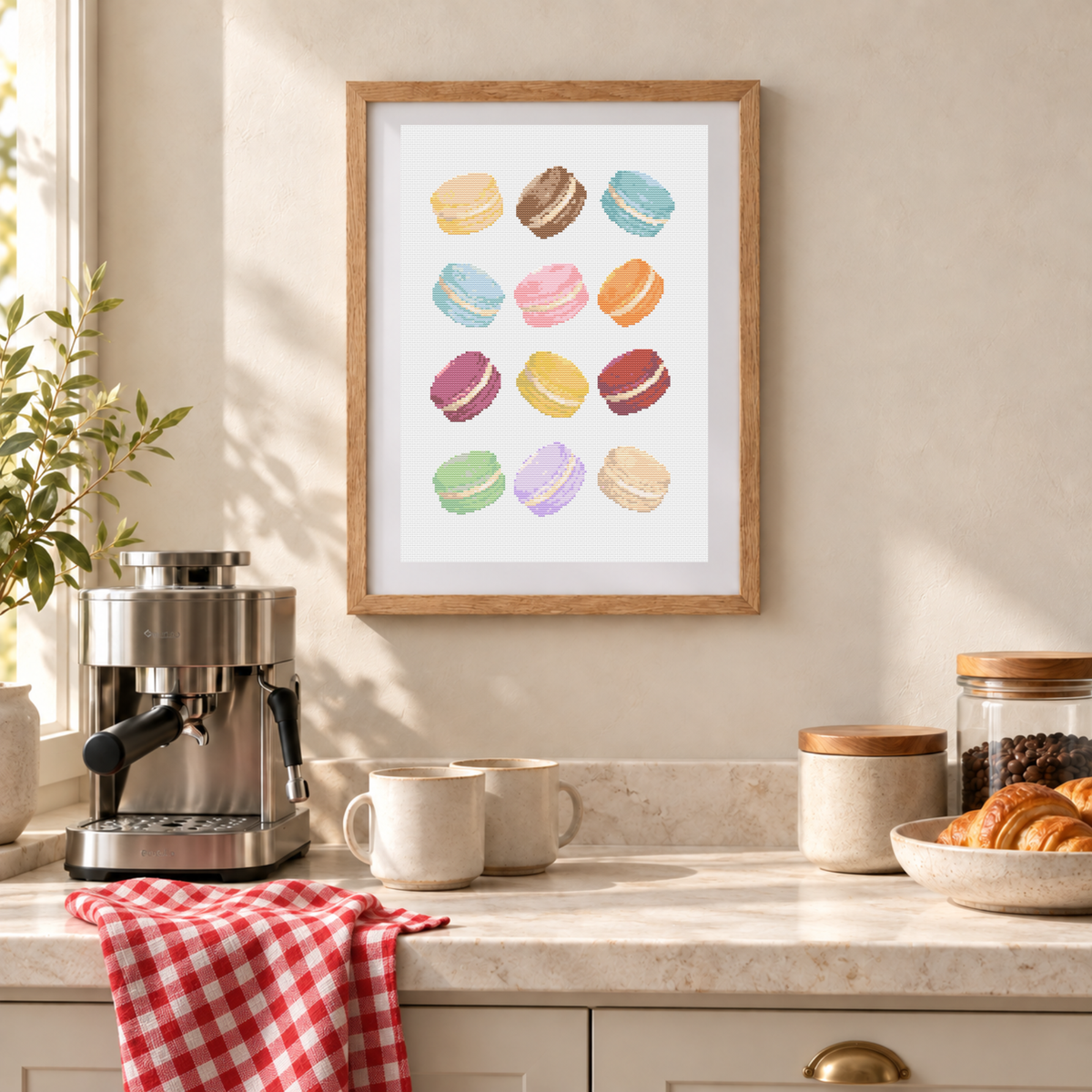 A framed cross-stitch pattern featuring a colorful macarons, displayed on a beige wall above a kitchen counter with an espresso machine, mugs, and pastries.