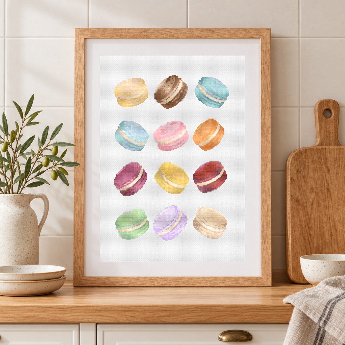 A framed cross-stitch patterns featuring a grid of colorful macarons on a white background, styled in a warm kitchen setting with a wooden countertop, tiled wall, a potted plant, and a wooden cutting board.