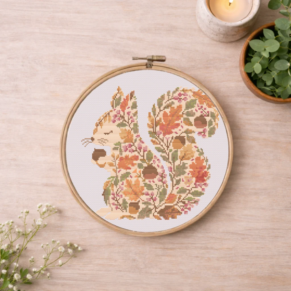 A circular embroidery hoop on a light wood surface, featuring a stitched squirrel design filled with autumn leaves and acorns in warm orange, brown, green, and pink tones. The hoop is styled with a candle, greenery, and small white flowers on a pale wood background.