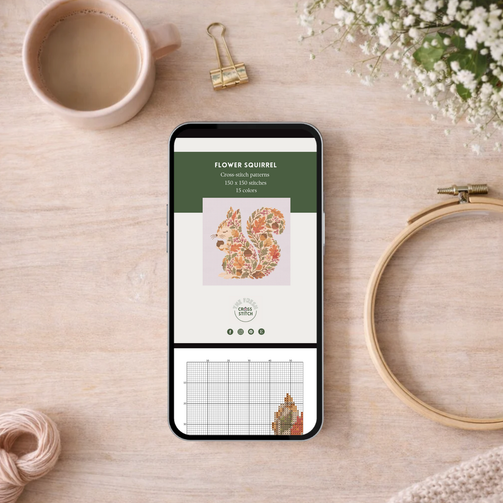 A smartphone lying on a light wood surface displays a floral squirrel cross-stitch pattern screen. The phone is surrounded by styled flat-lay props including a pink cup of coffee, a gold binder clip, white baby's breath flowers, a wooden embroidery hoop, and a ball of thread on a beige wood background.