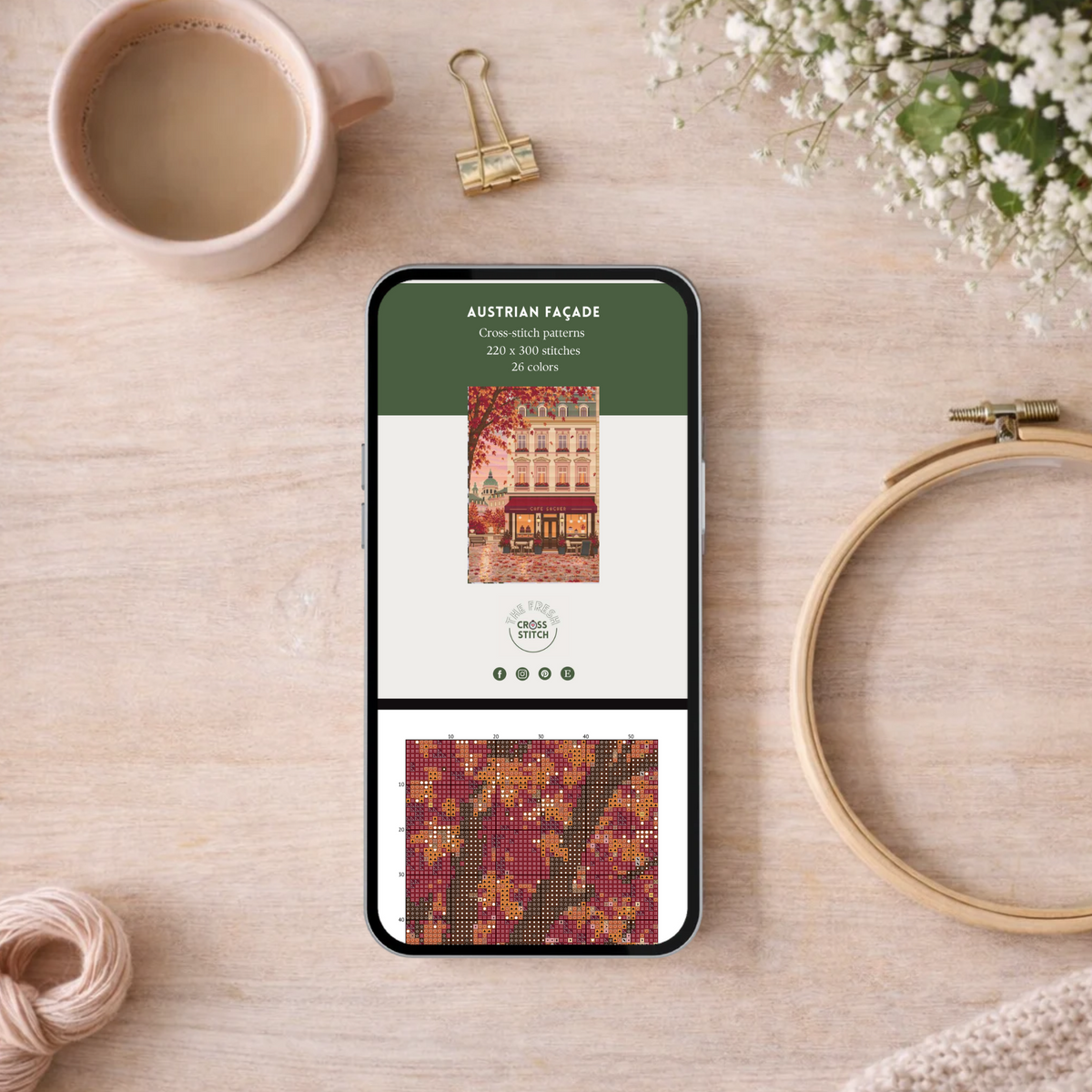 A smartphone lying on a light wood surface displays a cross-stitch pattern preview titled "AUSTRIAN FÀDAÇE." The scene is styled as a flat lay with a pink coffee cup, gold binder clips, white flowers, a wooden embroidery hoop, thread, and a soft knit fabric around it.