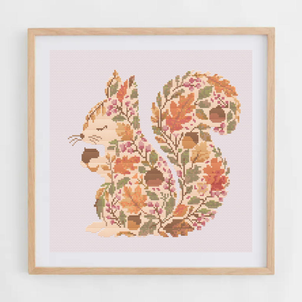 Framed cross-stitch art featuring a squirrel filled with autumn leaves and acorns, set against a pale pink background with a light wood frame.