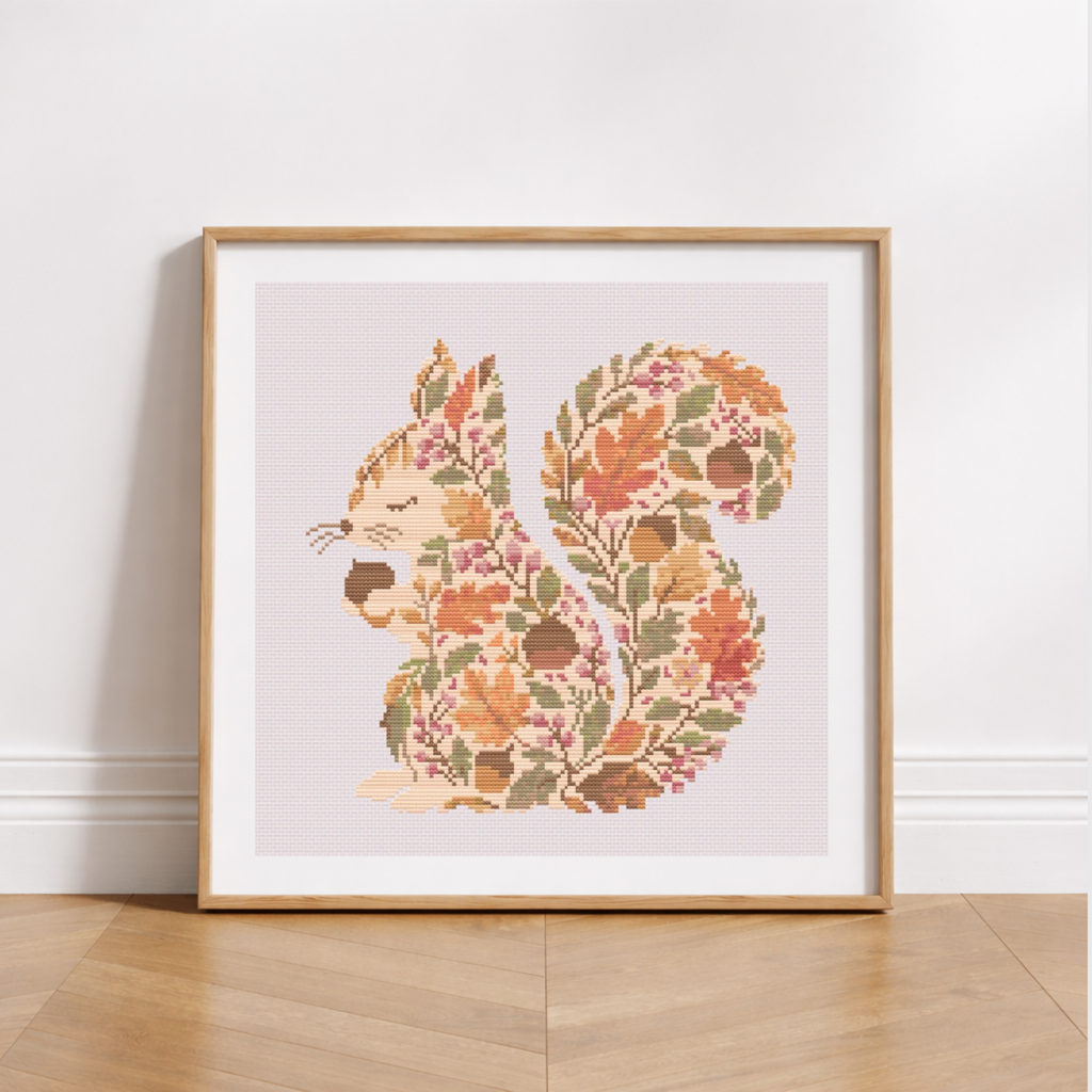 A framed cross-stitch or needlework artwork of a squirrel filled with autumn leaves and acorns, leaning against a white wall on a light wood floor.
