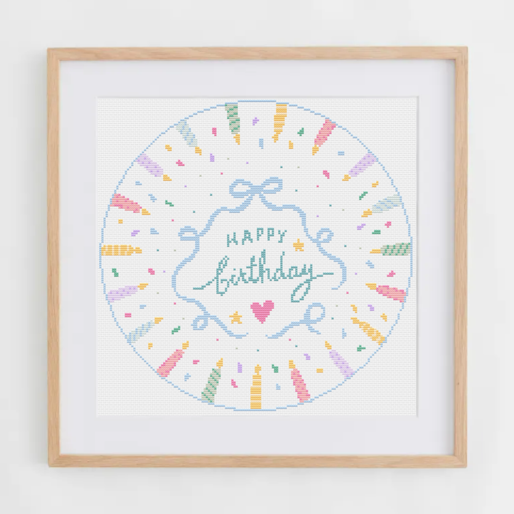 A framed circular cross-stitch design featuring a pastel 'Happy Birthday' script with colorful confetti and ribbon motifs, set in a light wood frame against a white wall.