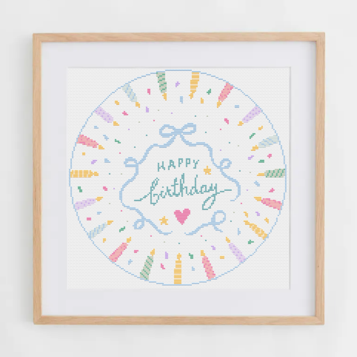 A framed circular cross-stitch design featuring a pastel 'Happy Birthday' script with colorful confetti and ribbon motifs, set in a light wood frame against a white wall.