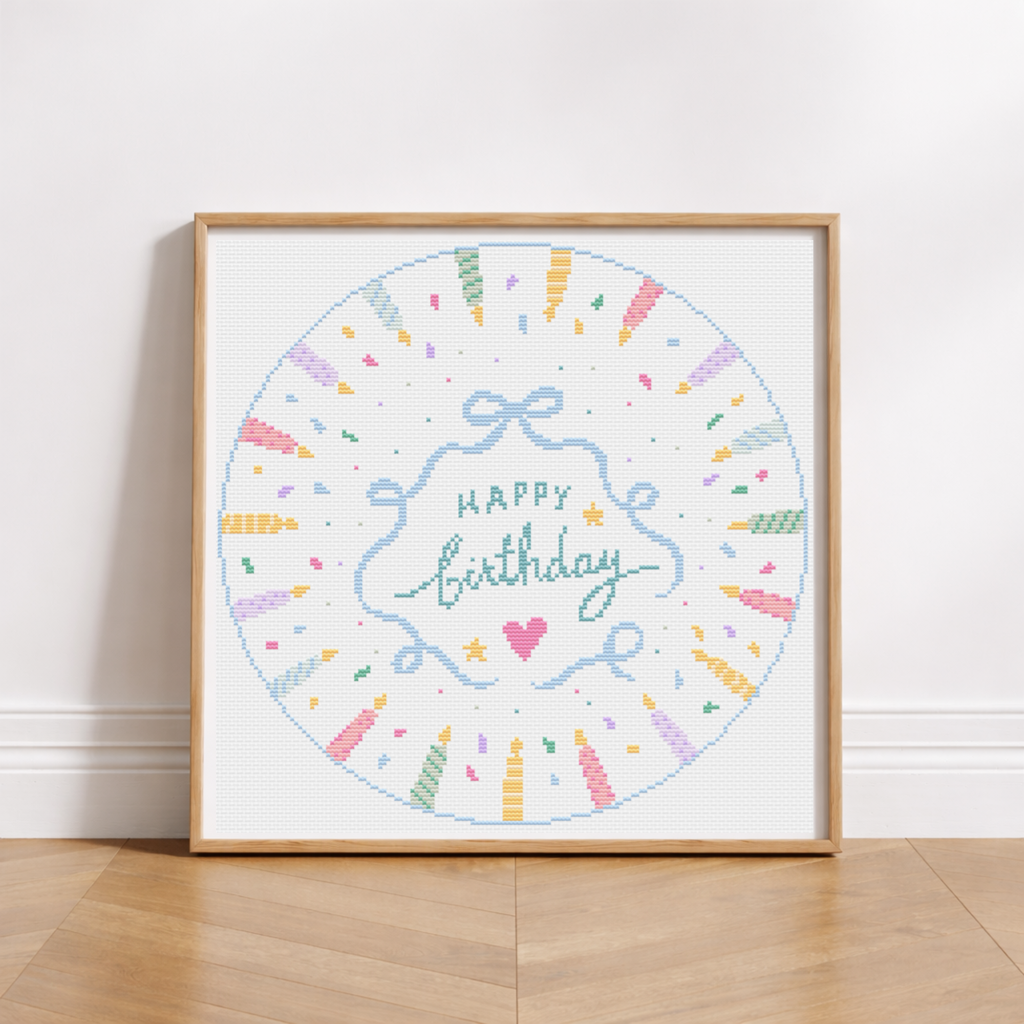 A framed circular cross-stitch design leaning against a white wall on a light wood floor. The artwork says “Happy Birthday” in blue script with a pink heart, surrounded by pastel confetti and candle motifs inside a light wood frame.
