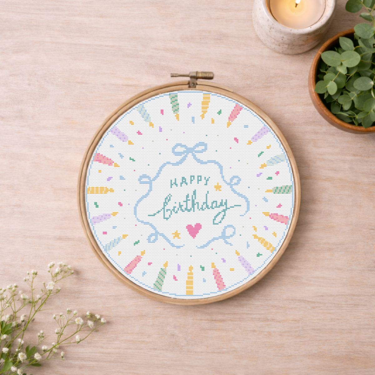 A circular embroidery hoop on a light wood surface, featuring a colorful cross-stitch design with the words “Happy Birthday” in the center, surrounded by pastel confetti, bows, and decorative accents. 